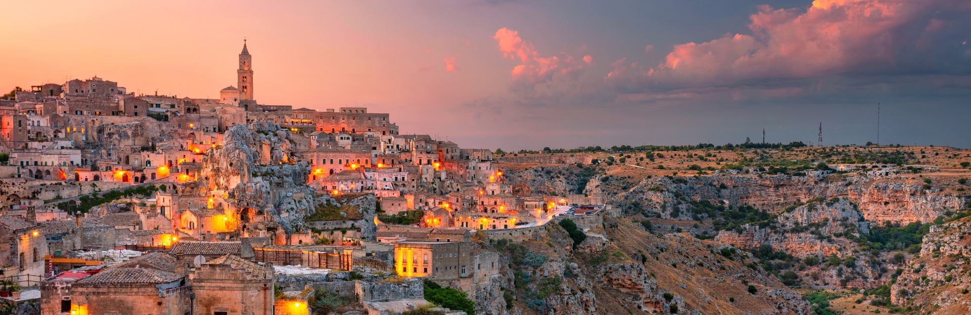 Paesaggio urbano storico al tramonto, luci calde, cielo drammatico, valle arida.