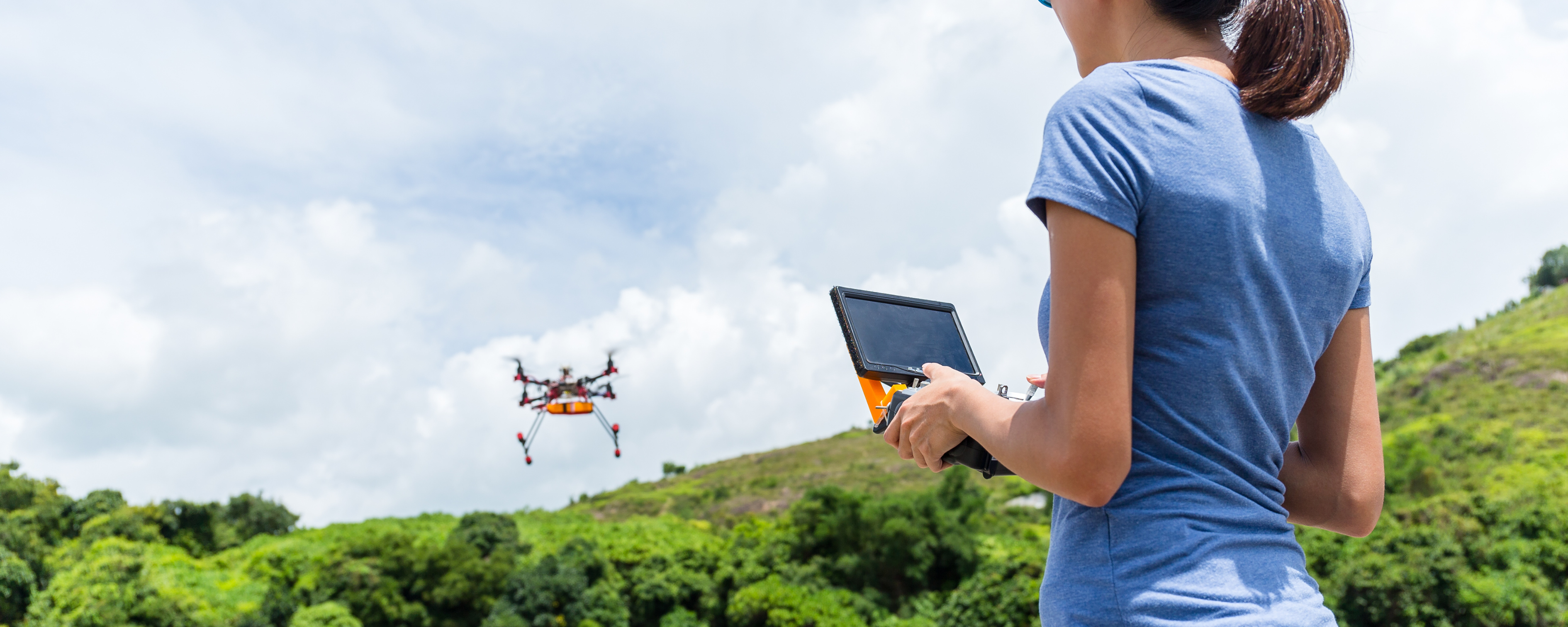 Donna pilota un drone arancione in un paesaggio collinare e verdeggiante sotto un cielo sereno.