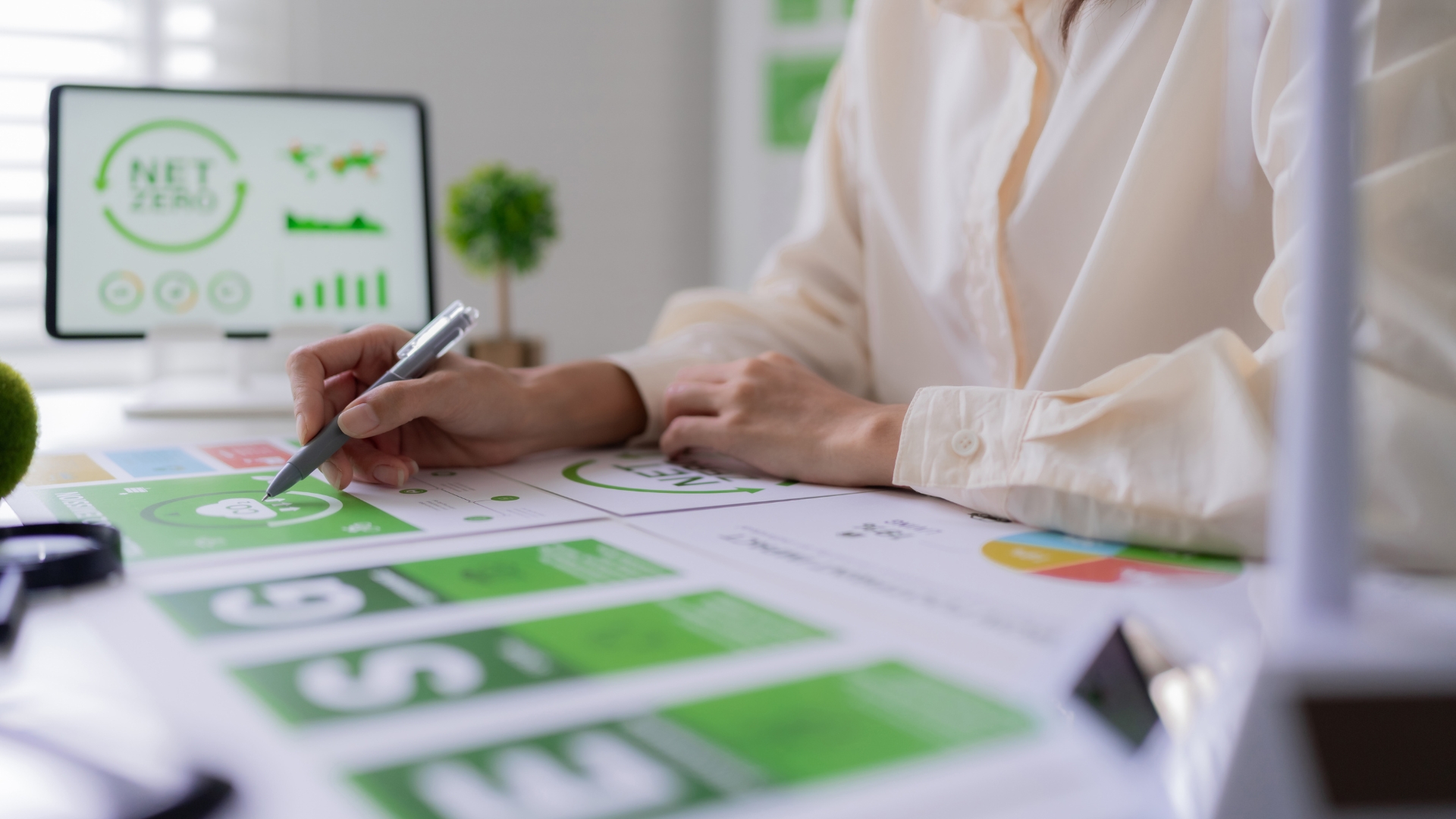 Persona che analizza grafici e statistiche su una scrivania, con focus su un documento con grafico "Net Zero" e uno schermo di computer che mostra dati sulla sostenibilità.